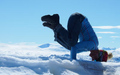 Kvinna står på händer och huvud i snö med blå himmel i bakgrunden.