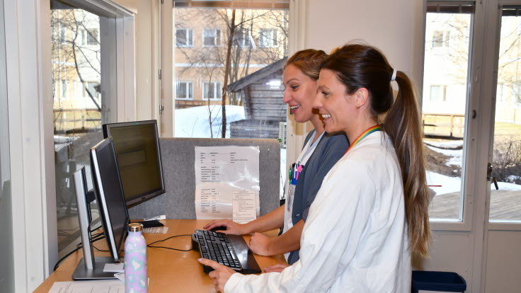 Enhetschef Antonia Lindgren och sjuksköterskan Tyra Eklund står på expeditionen vid ett skrivbord med två datorskärmar.